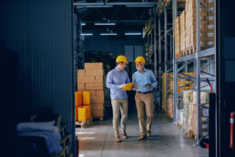 Two men in business casual outfits and hard hats walk through a warehouse as they converse over the details on a clipboard.